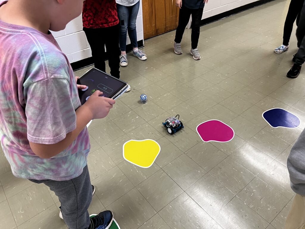 A student holds a tablet and maneuvers an mBot through colored shapes on a floor obstacle course, as other students watch. 