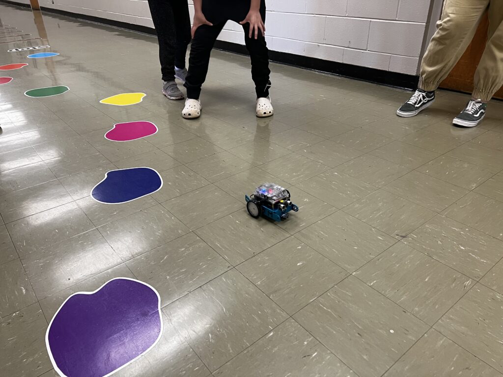 An mBot is traveling along an obstacle course of colored shapes in a hallway, with student feet standing around observing. 