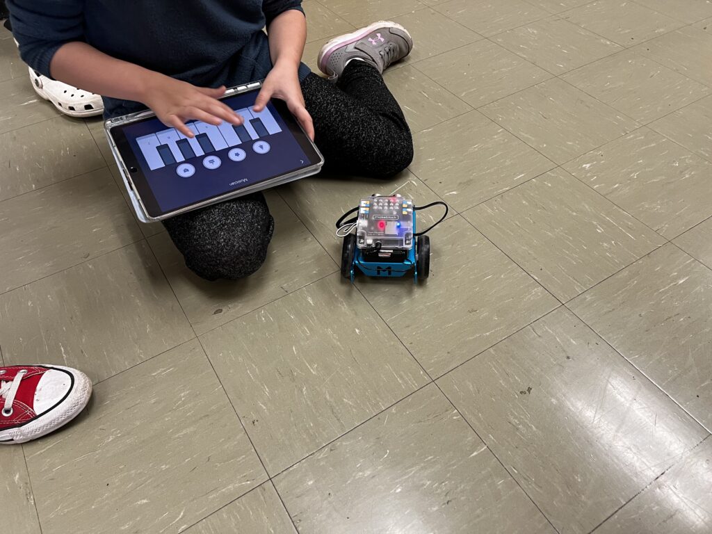 A student sits on the floor, using a tablet to move an mBot.