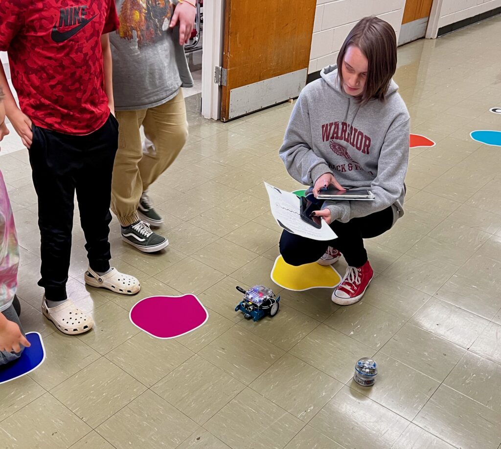 An individual crouches on the floor, holding papers and a tablet, explaining an mBot function to students standing nearby. 