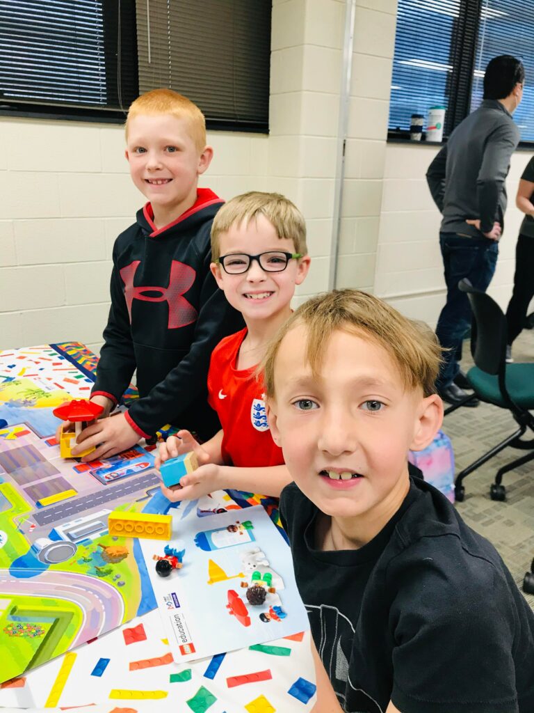 Three students sit smiling at a table, working on a Lego build. 