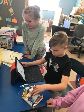 A student sits at a classroom table on a laptop, programming a Lego, with another student looking over their shoulder.