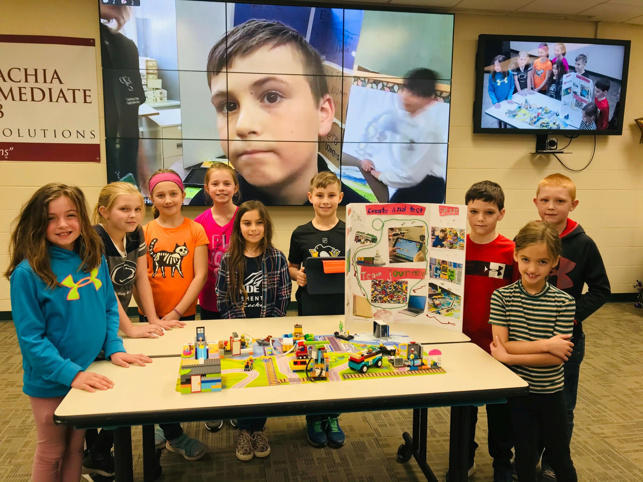 Students at a competition gather around a table with legos and a display board. Behind them, a large screen displays students virtually.