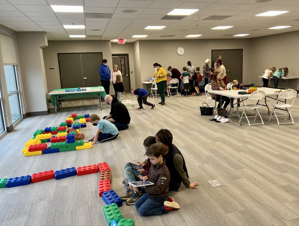 A large room is filled with groups of students surrounding different activities. Large LEGOs sit on the floor at certain centers, with tables of activities in the background. 