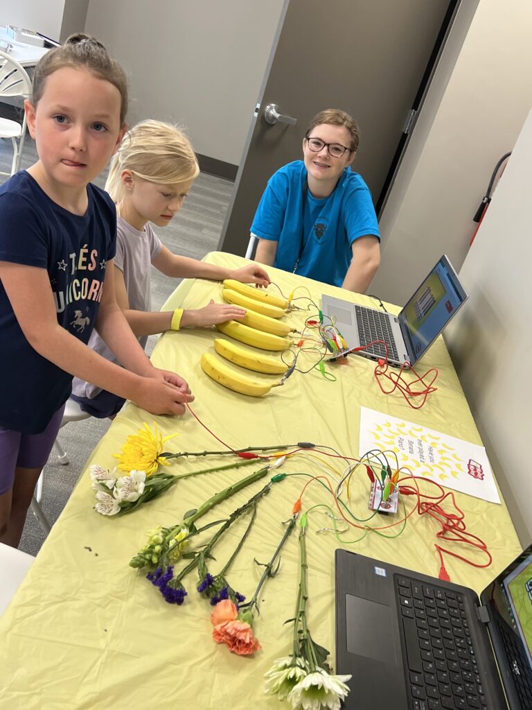 Two children get help with a banana piano: bananas hooked up to a laptop with Makey Makeys. 