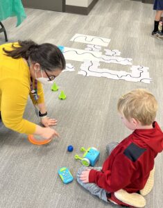 A child sits on the floor with a woman pointing at a blue robot. White maze outline pieces sit on the floor next to them. 