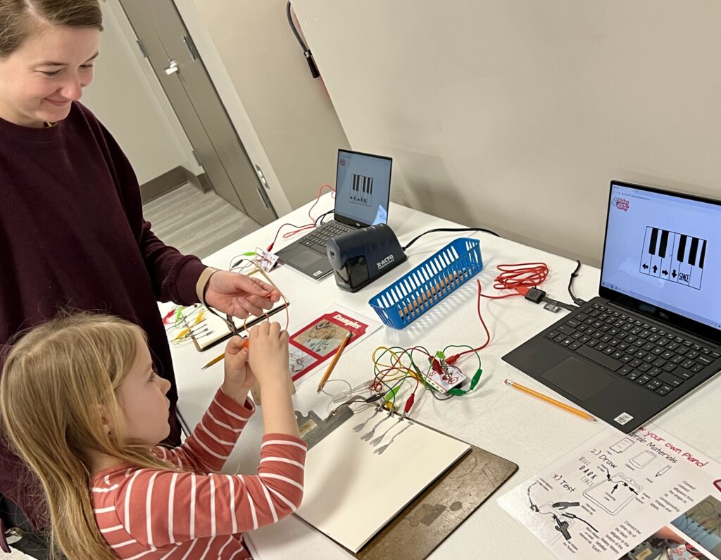 A child works on connecting Makey Makeys to a pencil drawing with the help of an adult. 