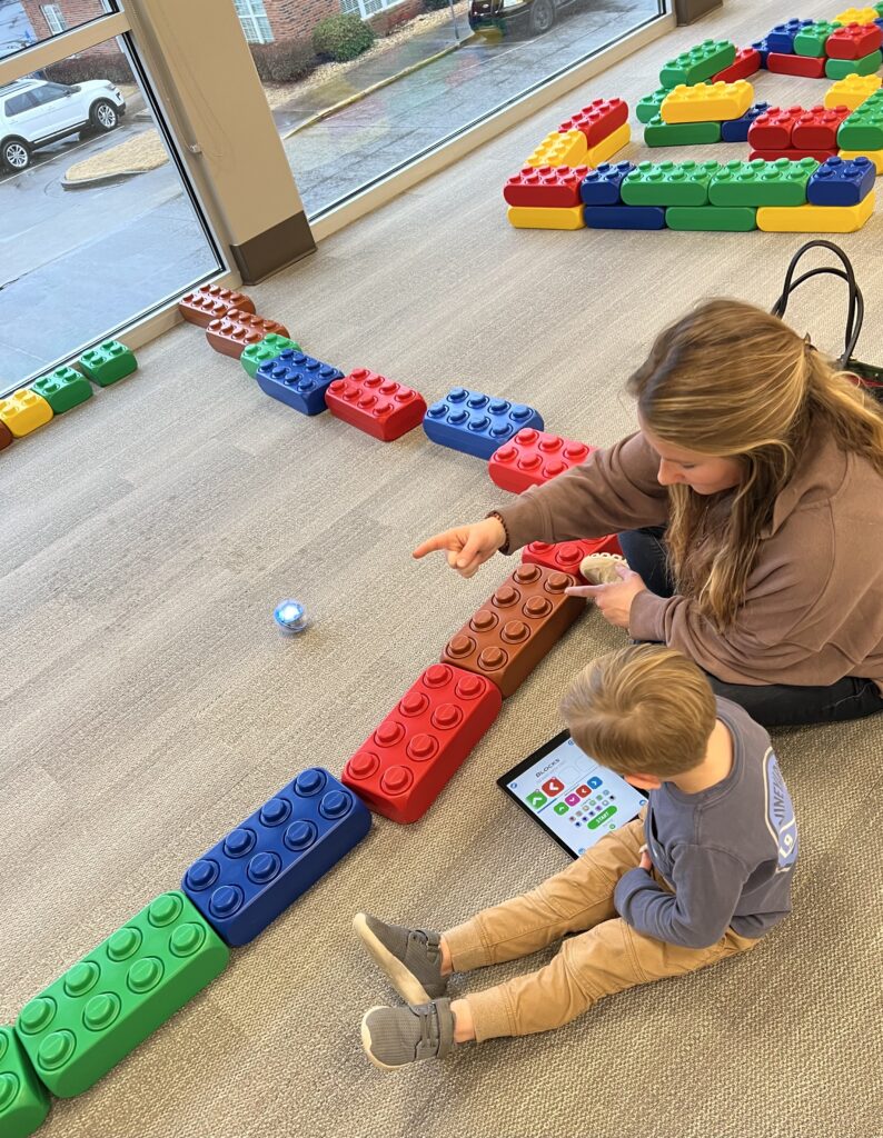 A child and adult sit on the floor next to a lego structure with a Sphero inside. The child has a tablet that is controlling it. 