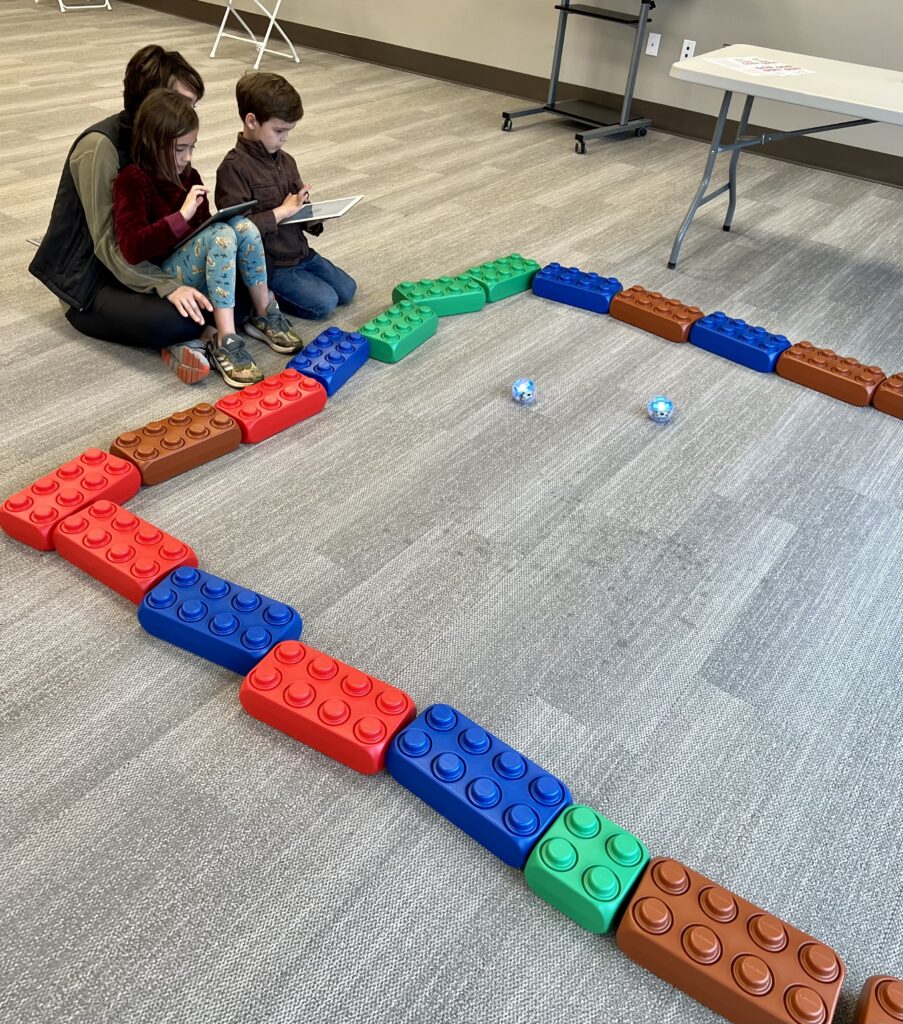 Two children sit with an adult on the floor, controlling two Spheros inside a Lego structure. 