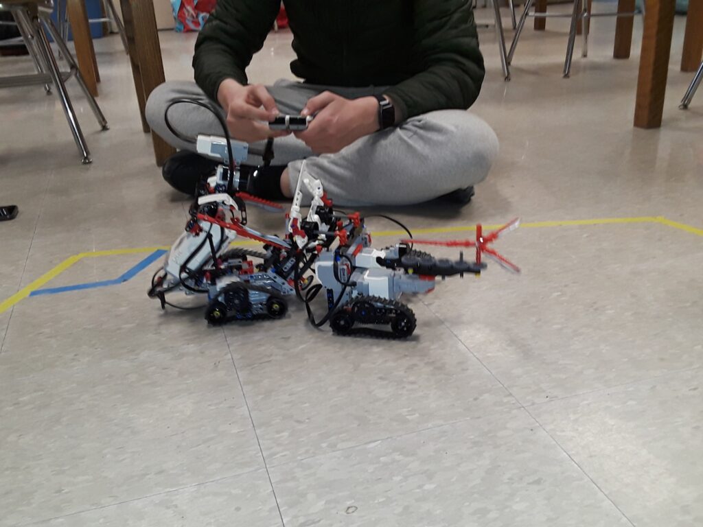 Two LEGO robots battle inside an arena on the floor made of tape. A student with a remote sits behind them. 