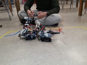 Two LEGO robots battle inside an arena on the floor made of tape. A student with a remote sits behind them.