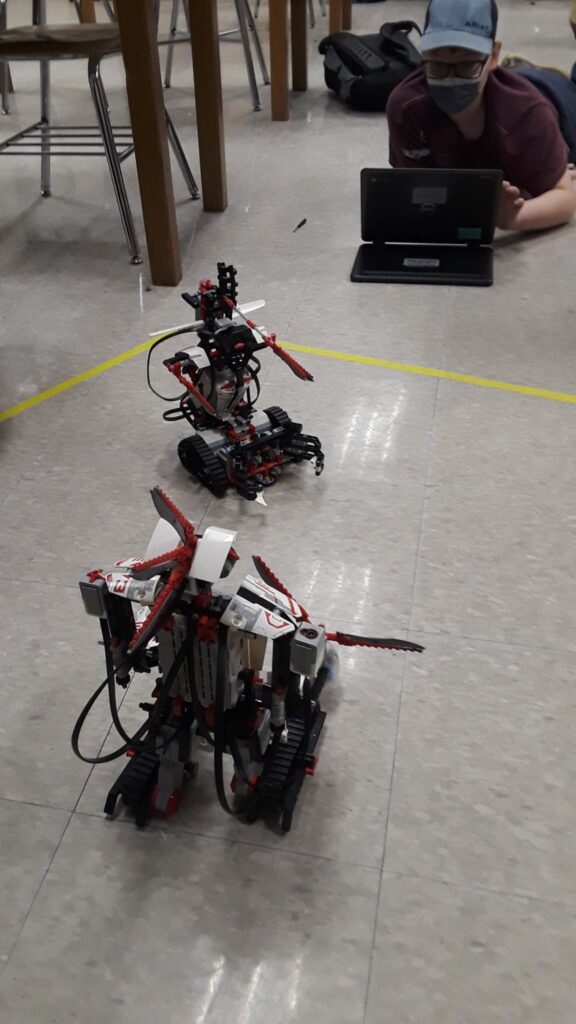 A student lies on the floor with a laptop, looking on to two battling LEGO Robots. 