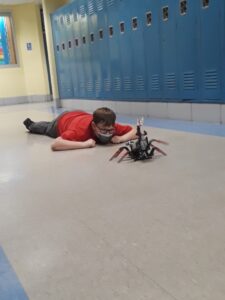 A student lies on the hallway floor, looking at a robot creation. 