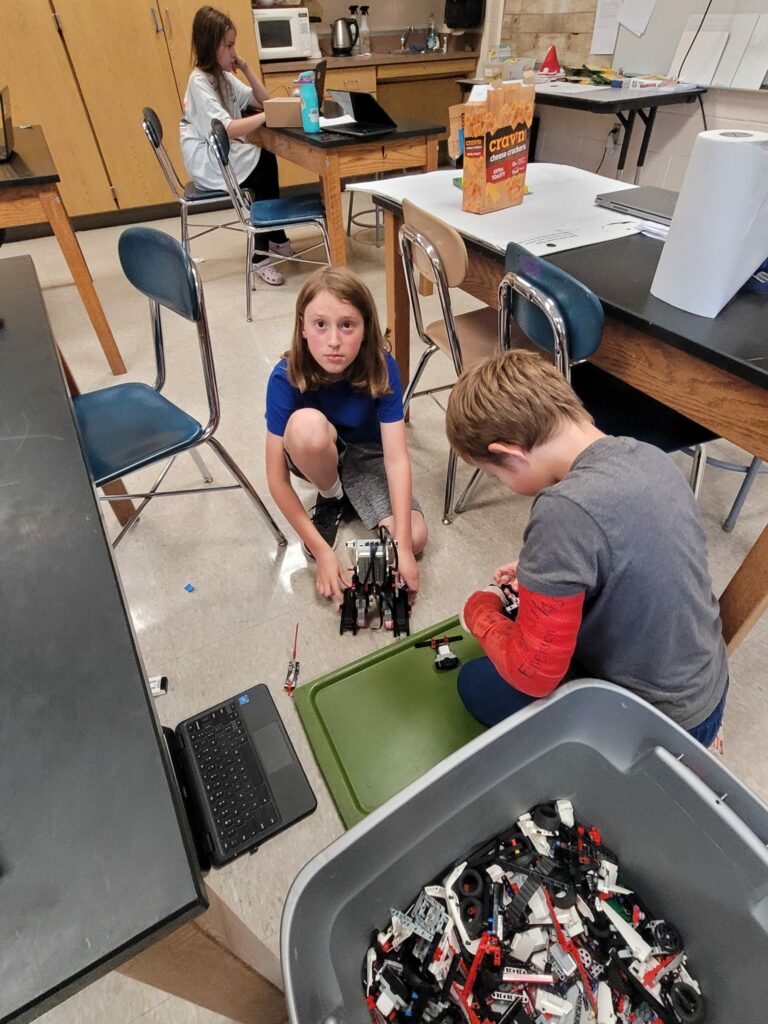 Students sit on the floor with a laptop and a bin full of LEGO robot pieces, building a creation together. 