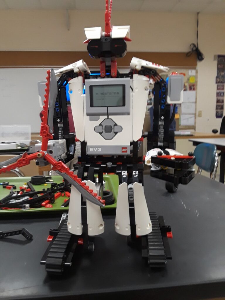 A white and red LEGO Robot with arms, legs, and a face stands up tall on a classroom table. 