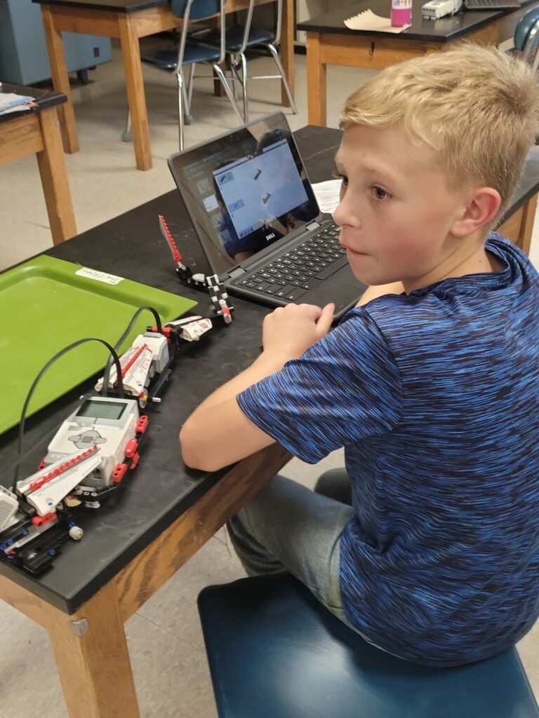 A student sits at a classroom table, programming a robot on a laptop. 