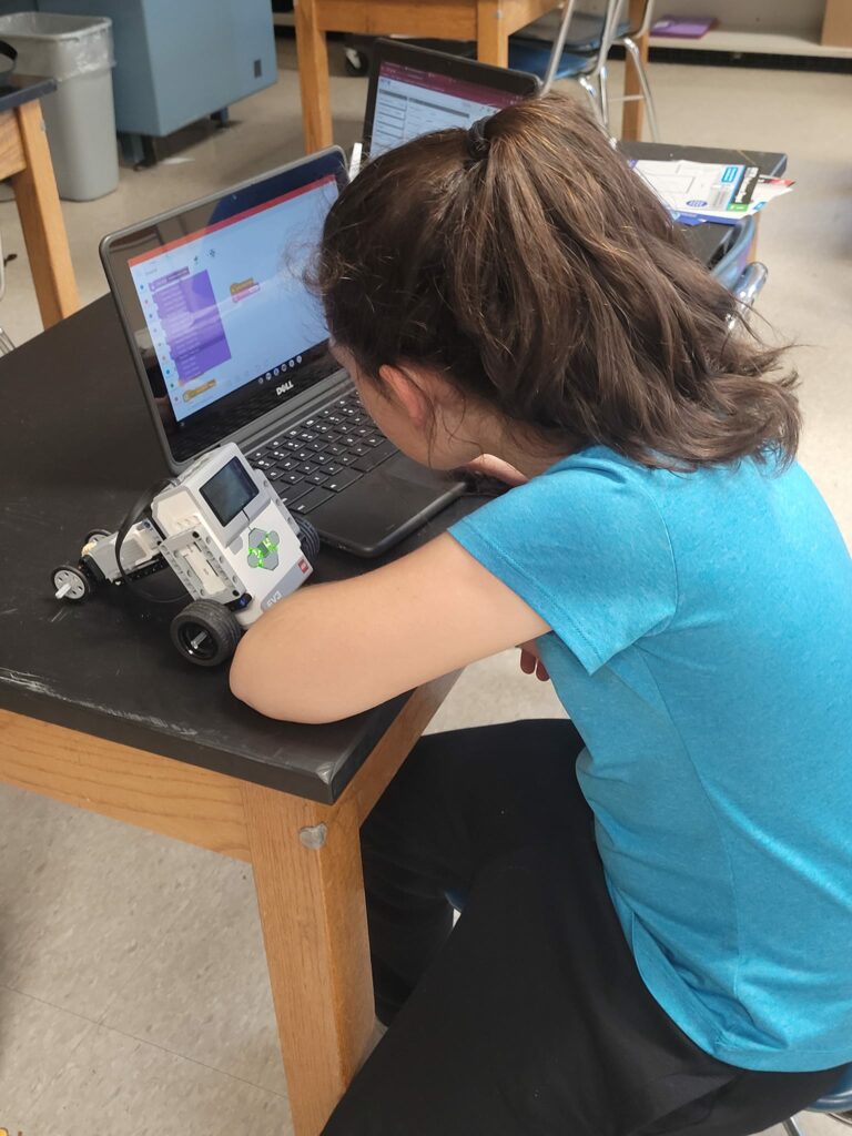 A student sits at a laptop, programming a robot. 