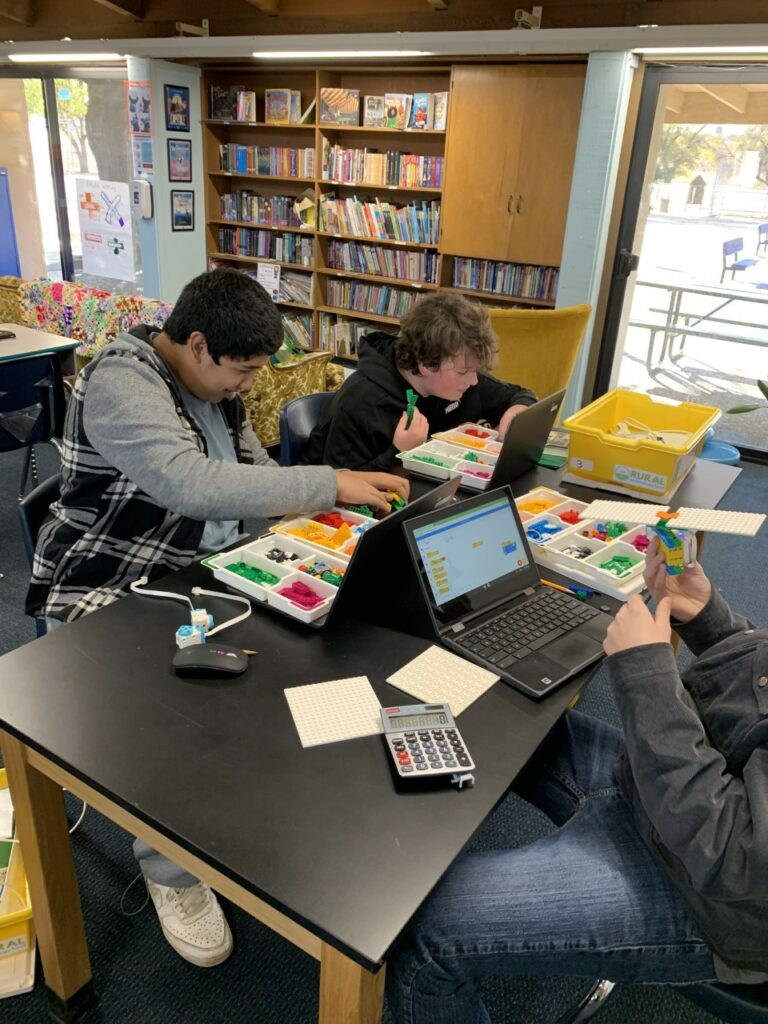 Three students sit at a classroom table, working on LEGO Spikes and laptops. 