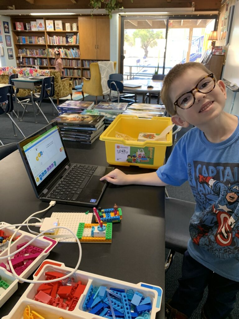 A smiling student sits at a table working with a LEGO Spike and a laptop. 