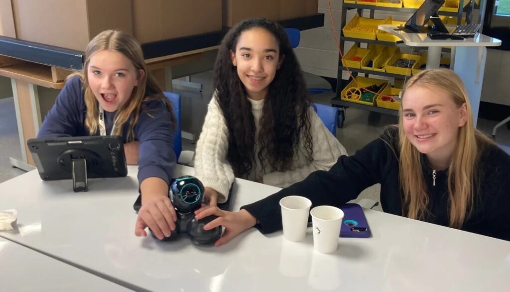 Three students sit at a classroom table, smiling with their hands together on a bot.