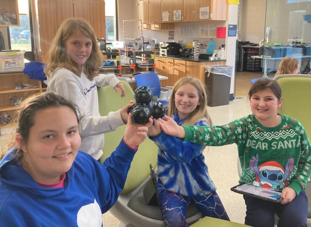Four students hold a bot, smiling while sitting in a circle. They appear in a classroom full of various STEM equipment. 