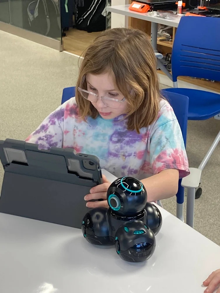 A student works on an ipad, programming a bot sitting on the classroom table. 