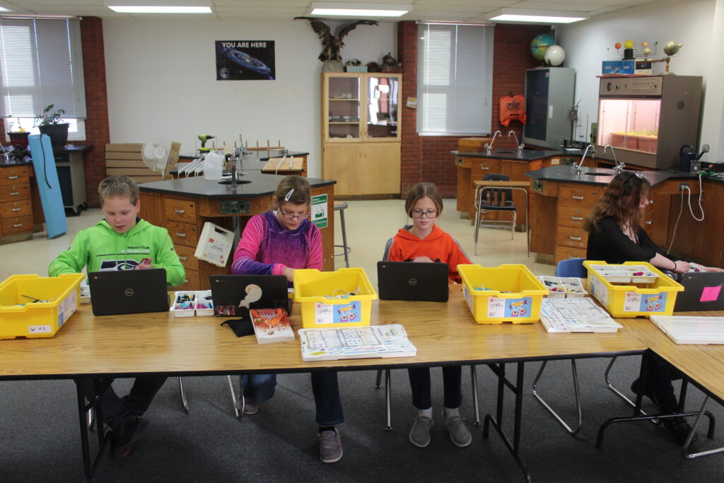 Four students sit at a long, L-shaped table, each with a Lego SPIKE kit and a laptop. 