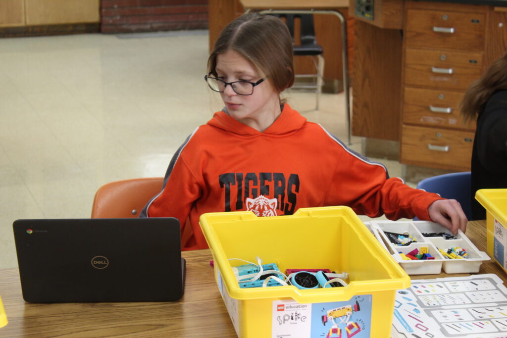 A student sits at a classroom table, looking at a laptop while reaching for Lego SPIKE pieces. 