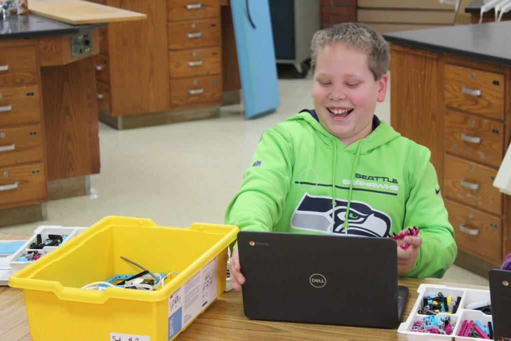A student sits beaming at a classroom table with a laptop and a Lego SPIKE kit. 