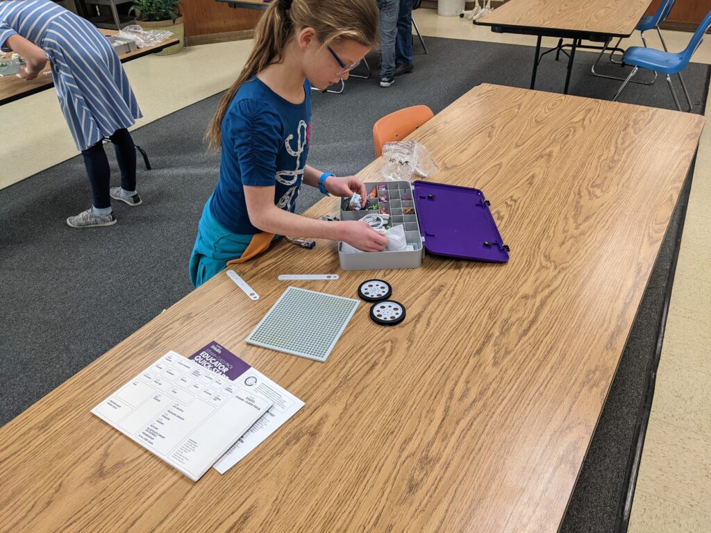 A student stands at a classroom table, sorting through a box of Little Bits. 