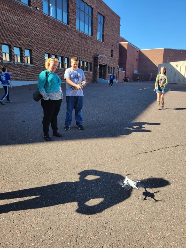 A low-flying drone is hovering above the cement while a student holds the remote. 