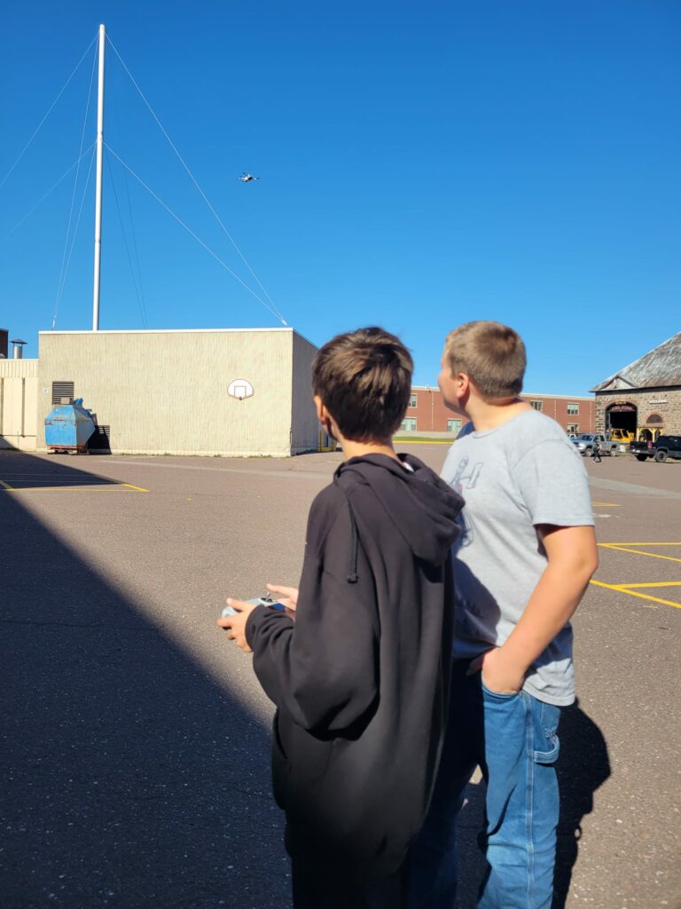 Two students stand outside of a school building, operating a drone. 