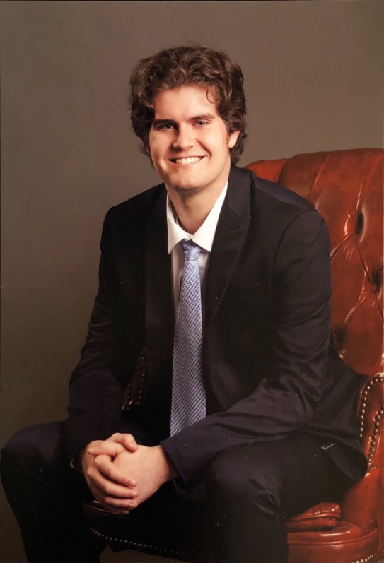 Headshot of Gabriel in a suit, smiling and sitting in a leather chair. 