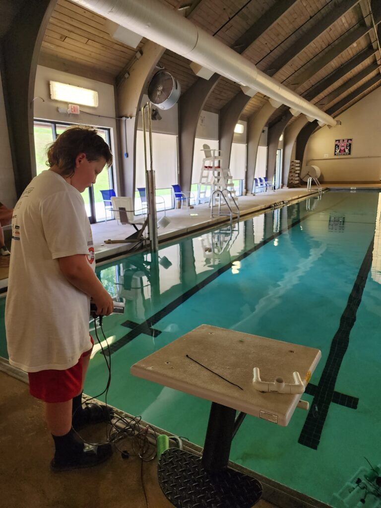 a camper stands next to the pool with a remote control, attached to a robot. 