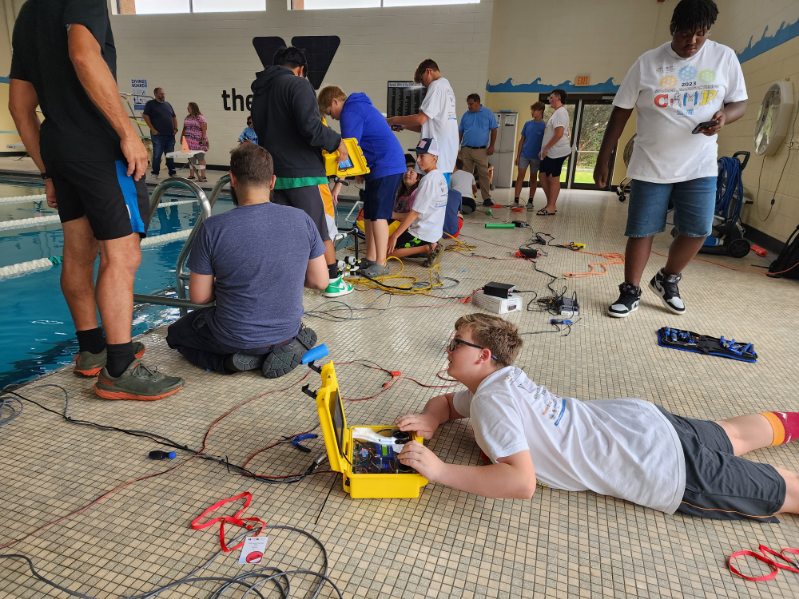 Many students gather around a pool, with one student lying on the ground, surrounded by equipment and wires. 