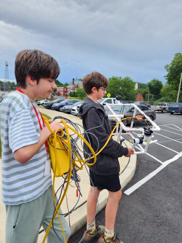 Two campers carry their robotics equipment and many wires through a parking lot.  
