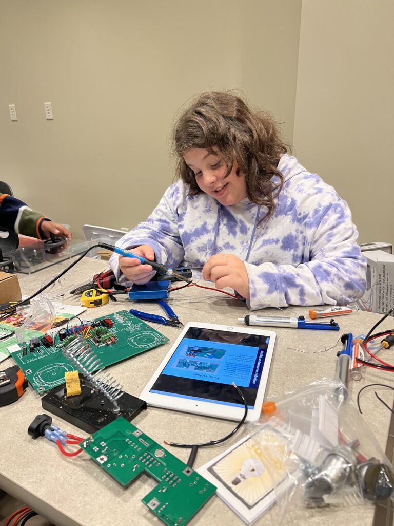 A student sits at a table with equipment and an iPad, smiling and constructing a piece of a robot. 