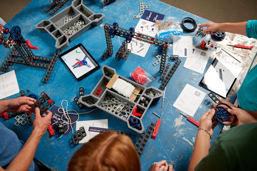 Parts of a Sphero Blueprint sit scattered on a table, with several individuals gathered around working wtih various pieces. 