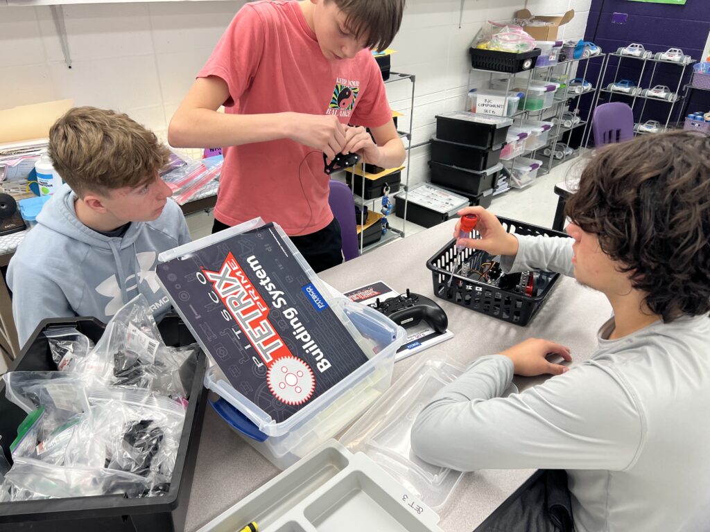 A student stands at a classroom table, working on a TETRIX Robot and controller with another student. 