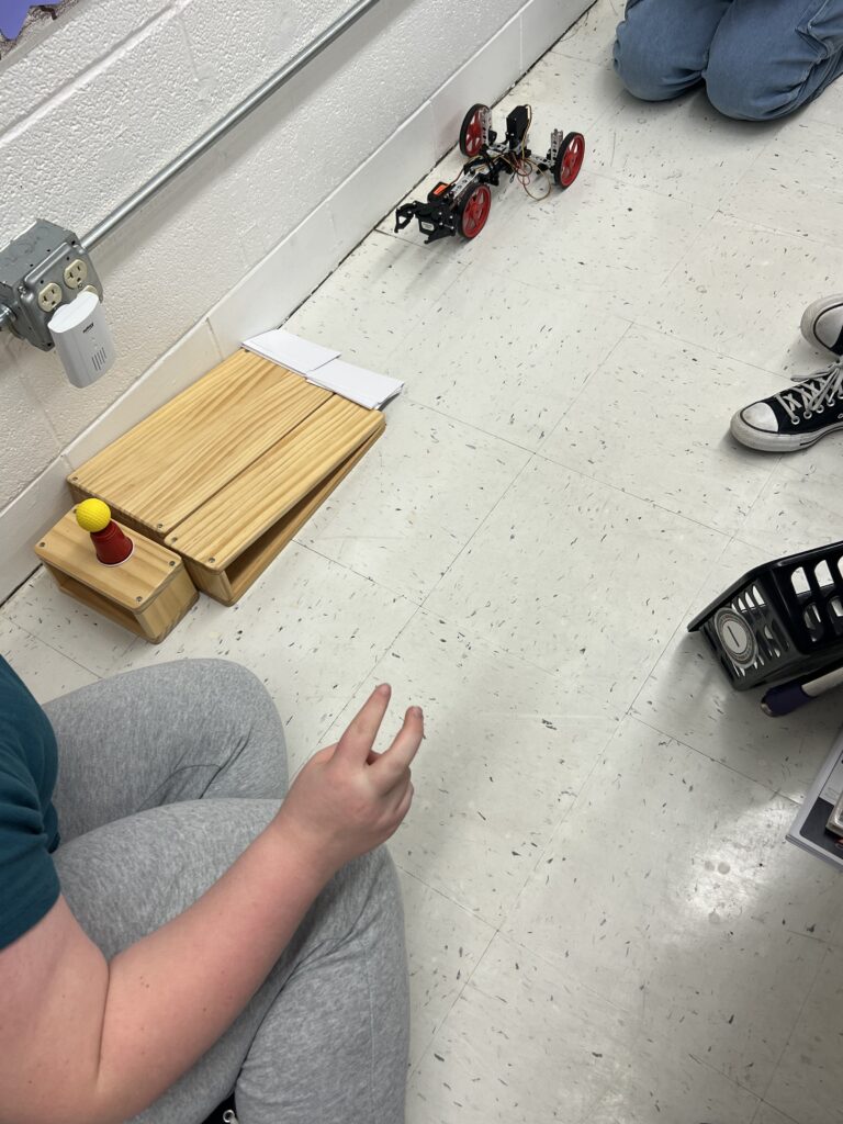 A TETRIX Robot is shown on the classroom floow, appearing ready to go up a ramp that has a target at the end. Students gather around the robot. 