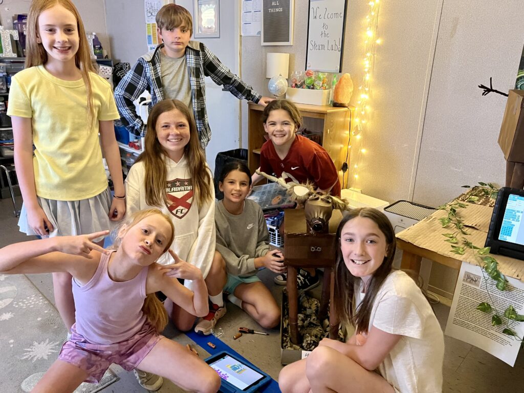 Students smile in a group, surrounding their robotics creation and supplies. 