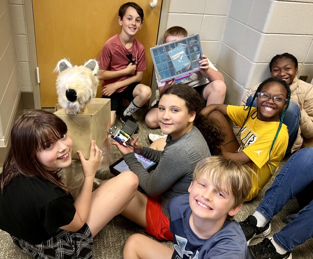 A group of students sit on the carpet of a classroom, smiling with their animal creation and robotics components. 