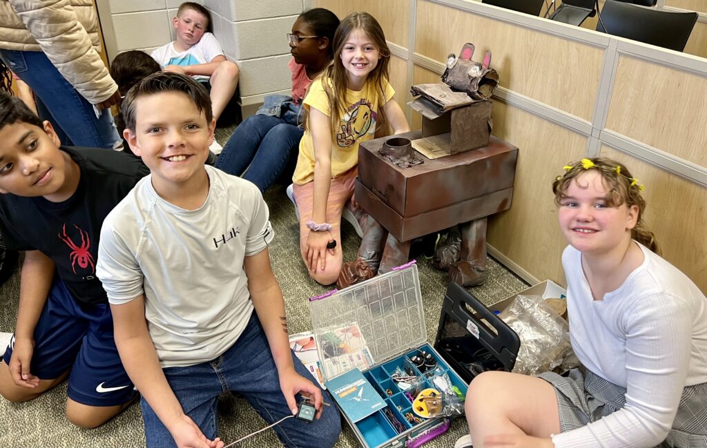 Students gather around their animal creation and an opened Hummingbird Robotics kit. 