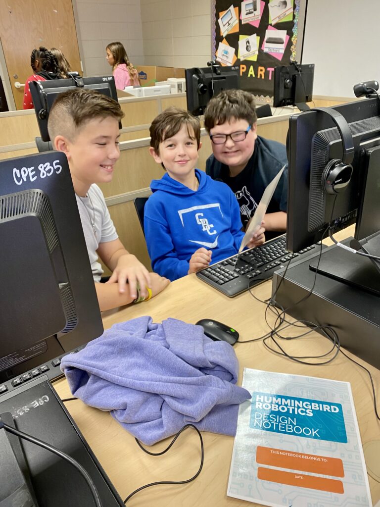 Three students sit at a table, working on a desktop computer.