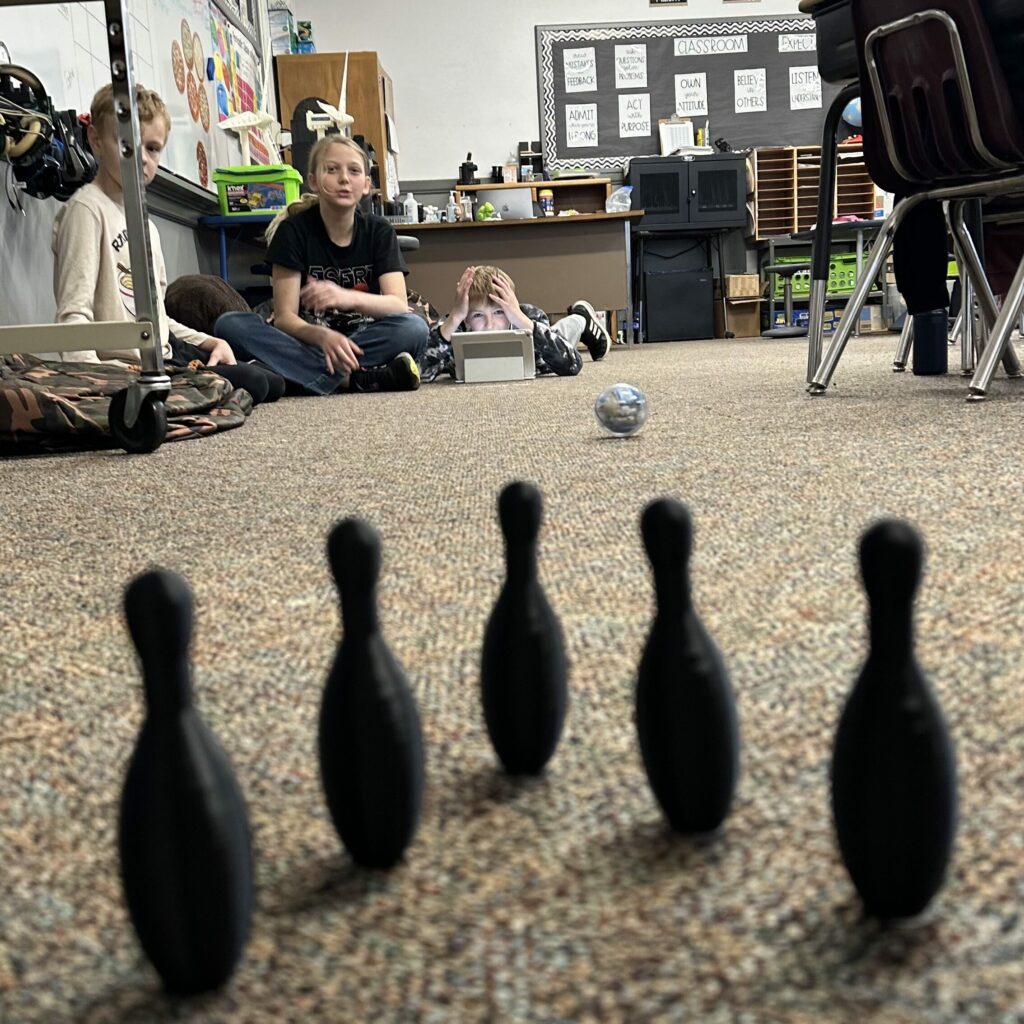 3D-printed bowling pins sit on a carpet, with students directing a Sphero Bolt to knock them over. 