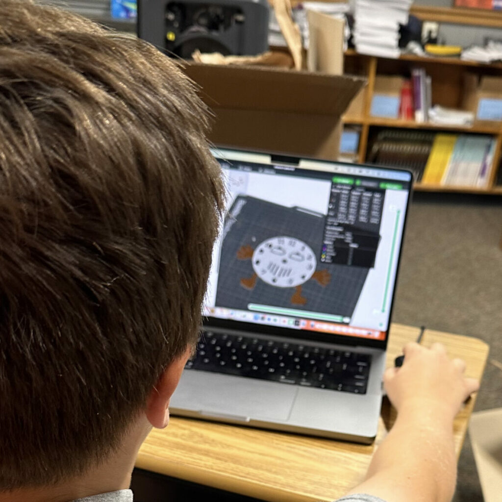 A student sits at a laptop and works on a 3D design of a smiling clock face. 