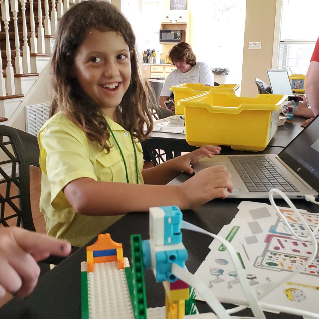 A student smiles brightly while working with a Chromebook and LEGO Spike creation. 