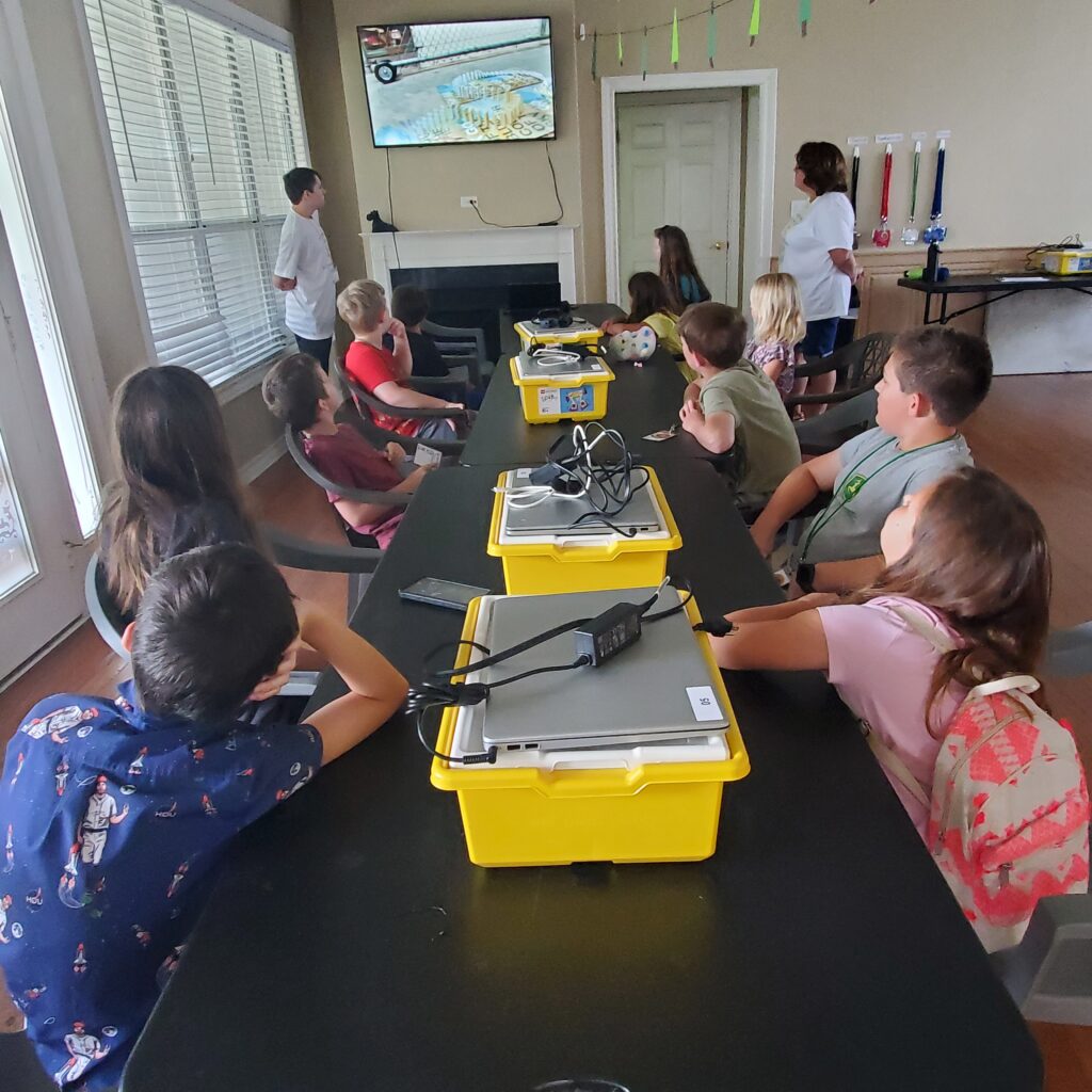 Students sit around a table wtih several LEGO kits and Chomebooks, listening to directions. 