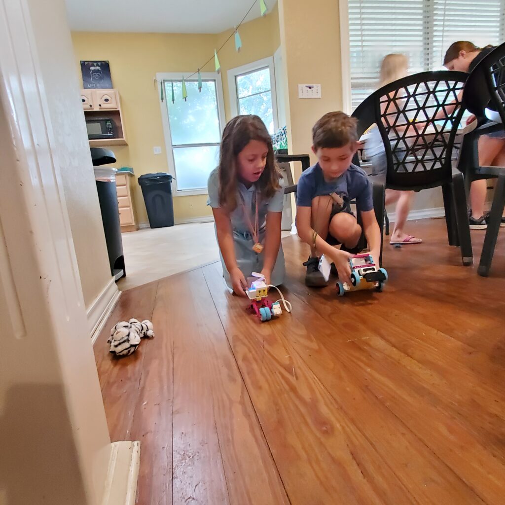 Two students kneel on the floor, setting up their LEGO Spike creations to test. 