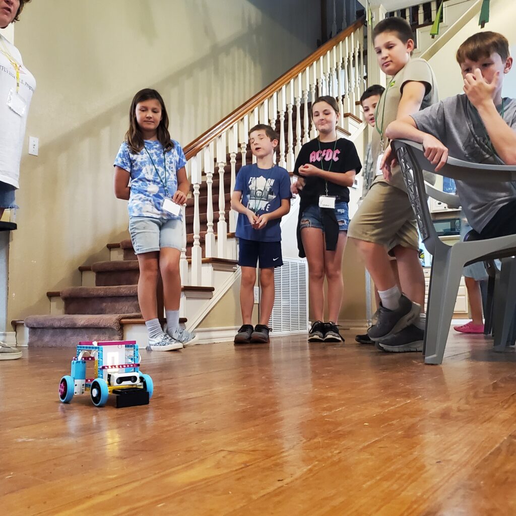 Students gather around a LEGO Spike robot, as it travels across the floor. 